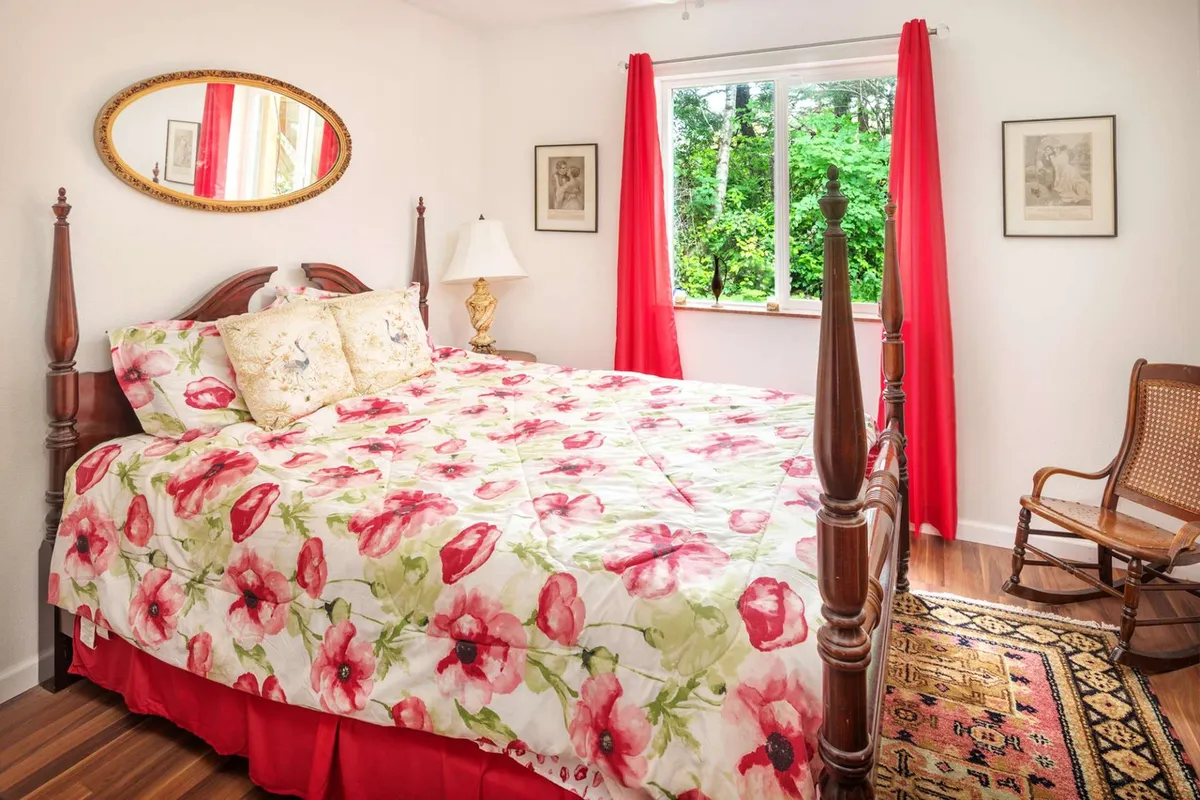 A bedroom featuring a floral-patterned bedspread, a large window offering lush greenery views, an ornate mirror, and a soft lamp. The overall atmosphere feels warm, welcoming, and perfect for restful sleep.