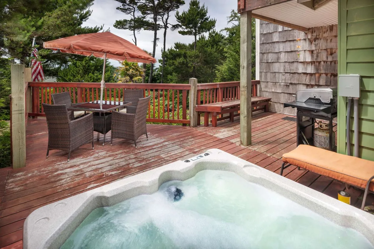 This exterior image features an expansive deck with a hot tub, outdoor dining table, umbrella, and grill, all surrounded by lush nature. It captures the seamless indoor-outdoor flow, offering a space for morning coffee, sunset cocktails, or relaxing soaks while enjoying scenic views.