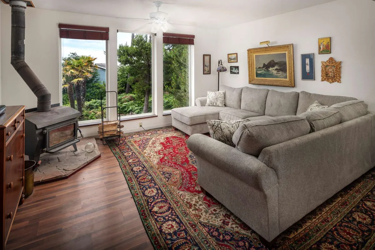 This interior image showcases a cozy living room complete with a comfortable sofa, wood stove, large windows, and thoughtfully placed artwork. The expansive windows flood the space with natural light and offer glimpses of the surrounding forest and possibly the ocean. The layout encourages both relaxation and social interaction, perfectly suited for family gatherings and group activities.