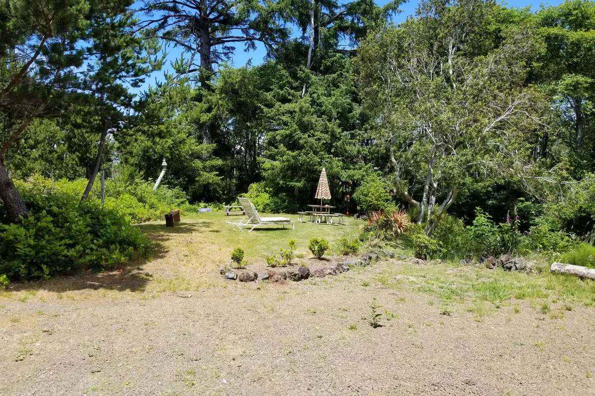An exterior seating area featuring two lounge chairs, a picnic table under an umbrella, and a surrounding of vibrant greenery. The calm setting evokes moments of relaxation and connection with nature, ideal for sipping morning coffee or enjoying a sunset together.