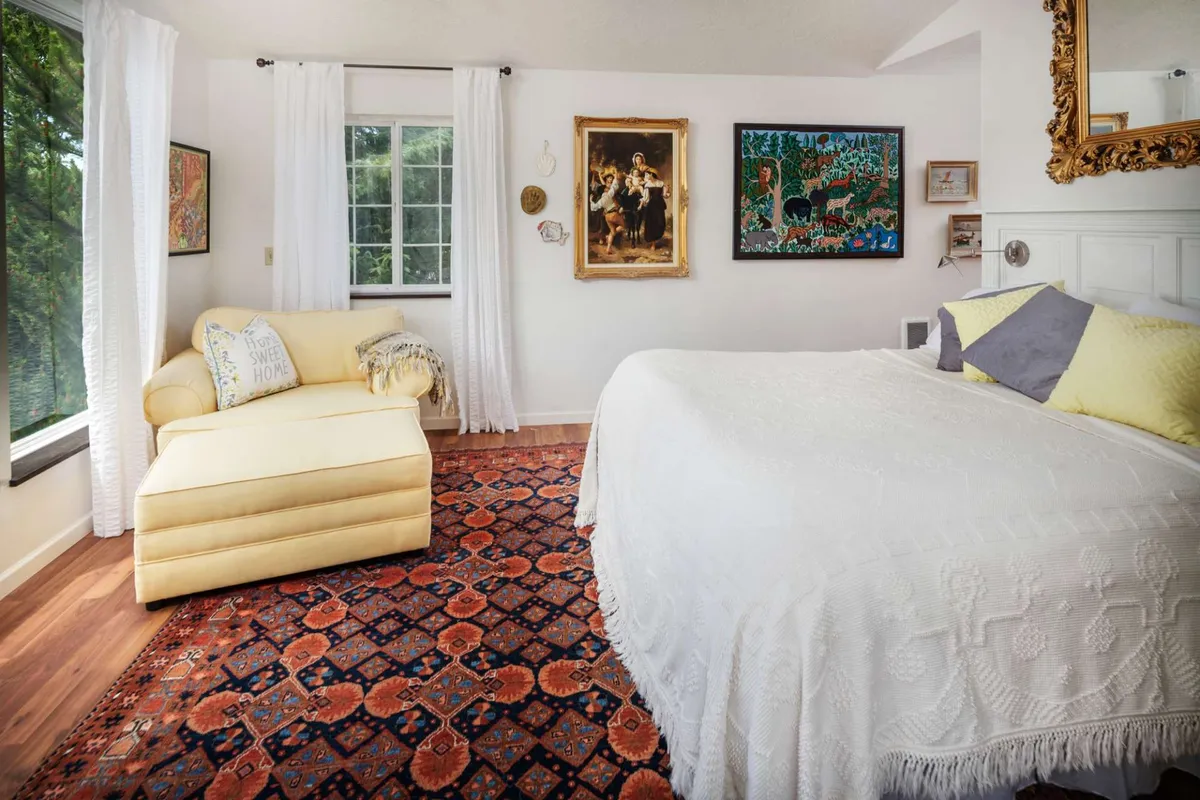 A serene bedroom with a large bed covered in a crisp white coverlet, a bright yellow chaise lounge near a window, colorful wall art, and abundant natural light streaming in. The space feels inviting and artistic.