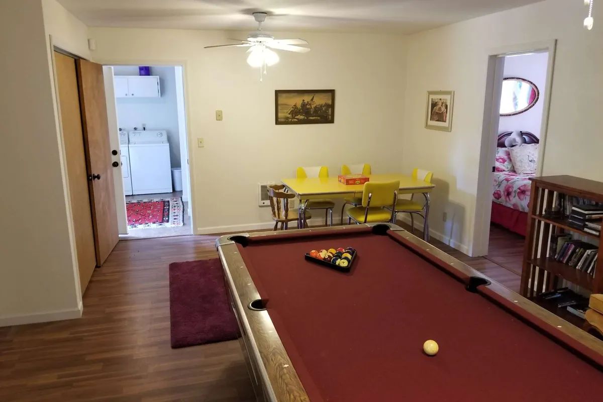 A cheerful game room featuring a pool table as its centerpiece, a vibrant yellow dining table off to the side, and a well-stocked bookshelf. The room is inviting, with natural light streaming in and space for both play and socializing.