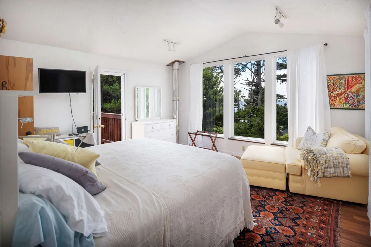 A tranquil bedroom with a large bed dressed in white bedding, spacious windows that let in natural light, a cozy seating area beside the window, and a decorative rug on the floor. The view outside the windows suggests privacy and calm, ideal for unwinding.