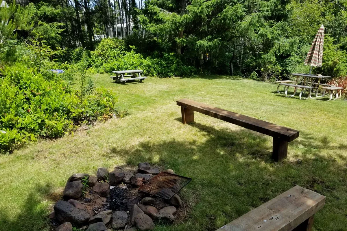 An inviting fire pit area surrounded by wooden benches and picnic tables, set within a backdrop of dense greenery. The space is designed for campfires, casual dining, and relaxed social gatherings, evoking warmth and camaraderie.