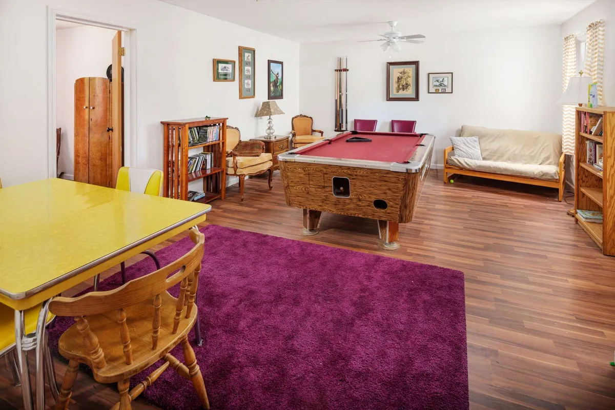 The bright, friendly game room features a pool table as its centerpiece, complemented by a yellow dining table, a purple rug, bookshelves, and a cozy seating area. The space is clearly designed for both active play and relaxed socializing.