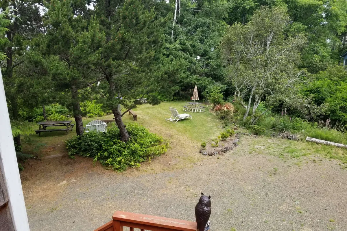 A peaceful garden scene featuring lounge chairs, a picnic table, a fire pit, and a decorative owl statue, surrounded by lush greenery. The area is set up for relaxing, dining, and gathering, with ample space for both people and pets, underscoring the inviting, communal outdoor atmosphere.
