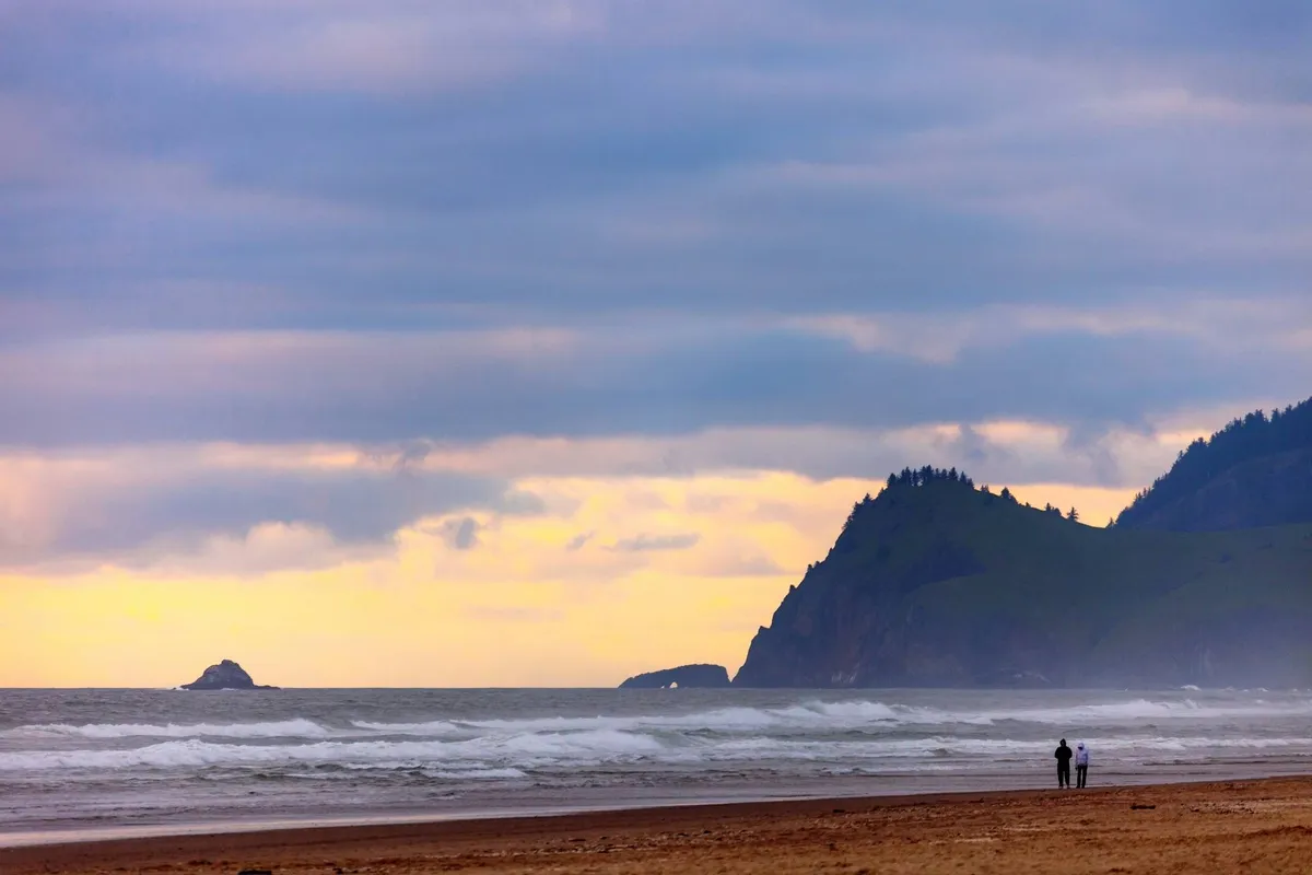 This coastal scene captures a couple walking along a sandy beach, with distant cliffs and waves crashing on the shore. The image conveys an inviting, wide-open beach ideal for sandcastle building, exploring, and family strolls.
