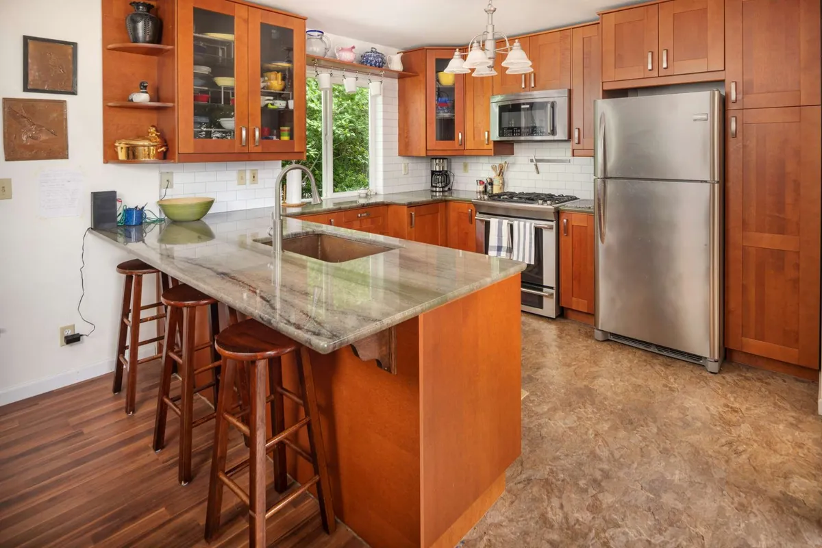 A stylish, modern kitchen with granite countertops, stainless steel appliances, open shelving with colorful dishes, and a large window with greenery outside. The setup is ideal for collaborative cooking, entertaining, and casual dining, with a visual connection to the outdoors.