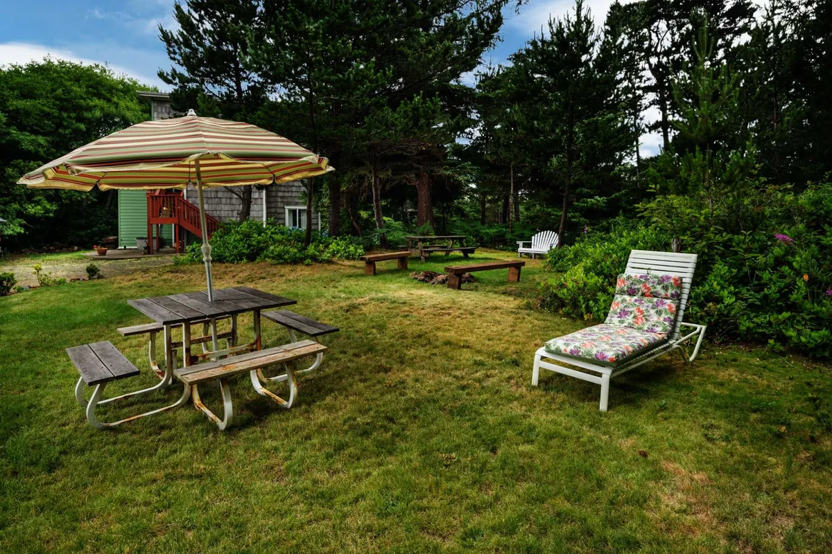 This image features an inviting outdoor area with a striped umbrella shading a picnic table, a comfortable lounge chair, and a fire pit, all set against a backdrop of greenery. The setup is clearly designed for socializing, dining, and relaxing outdoors, with the fire pit hinting at evening gatherings.