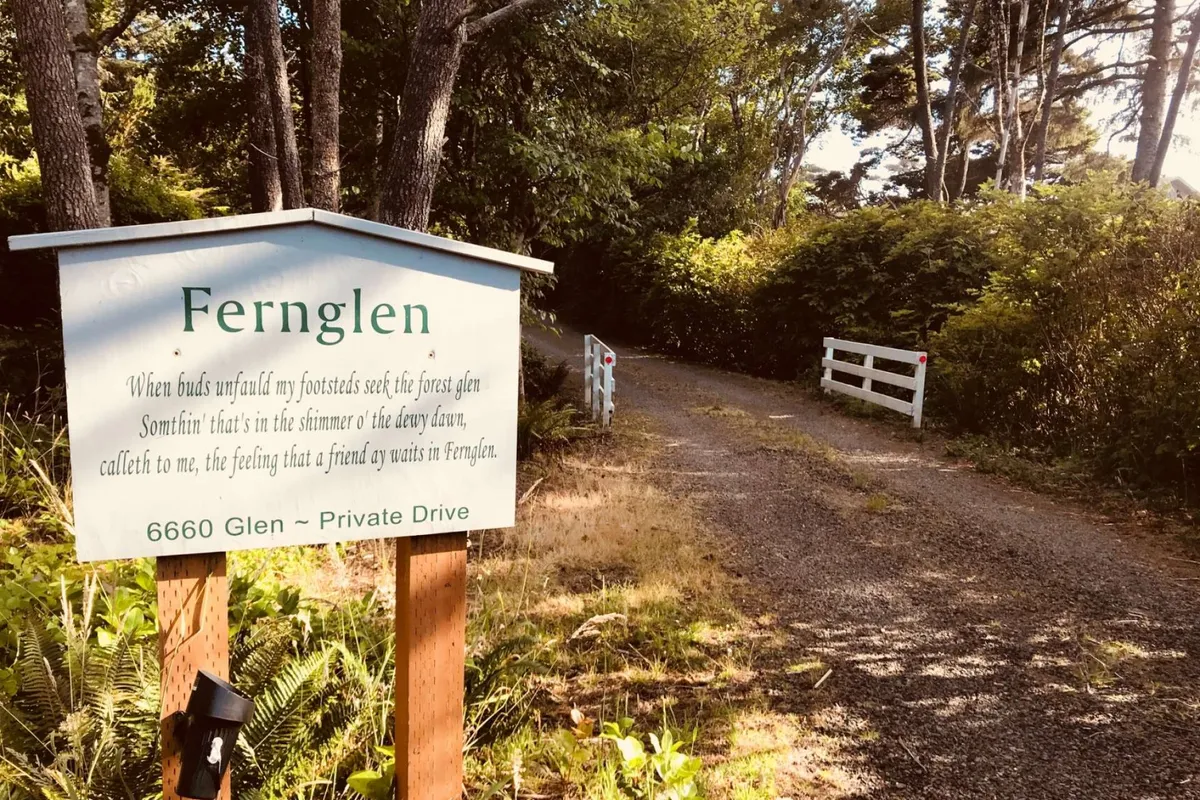 A serene entrance view with signage, a gravel path, and a gate, all bordered by tall trees. The setting exudes a sense of arrival and transition into a secluded woodland area, with dappled sunlight and inviting natural textures.