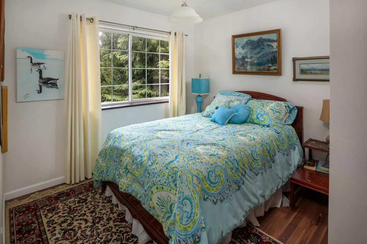 A serene bedroom with a full bed dressed in colorful bedding, accented by natural light from a large window and lively artwork on the walls. The ambiance is tranquil, offering a relaxing retreat after a day of fun.