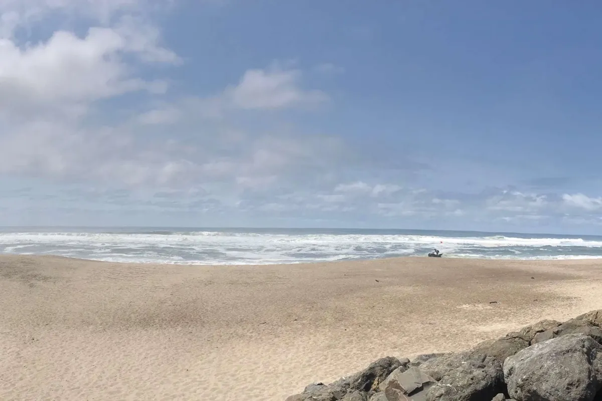 A serene sandy beach with ocean waves gently breaking along the shore, rocky outcroppings framing the scene, and a distant solitary figure walking. The tranquil atmosphere and natural beauty evoke the possibilities of shell collecting, beach walks, and sunset watching.