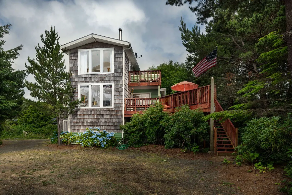 The hero image shows a spacious wooden deck bordered by vibrant hydrangeas and lush greenery, with large windows reflecting the forest surroundings. An American flag flutters gently atop the deck railing, hinting at warm gatherings. The inviting scene feels secluded yet open, perfect for outdoor relaxation and family moments.